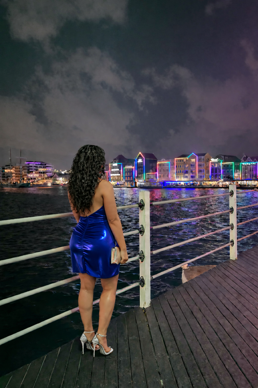 Surensca staat ’s avonds aan het water in Willemstad, Curaçao, met kleurrijke lichtreflecties op de achtergrond.