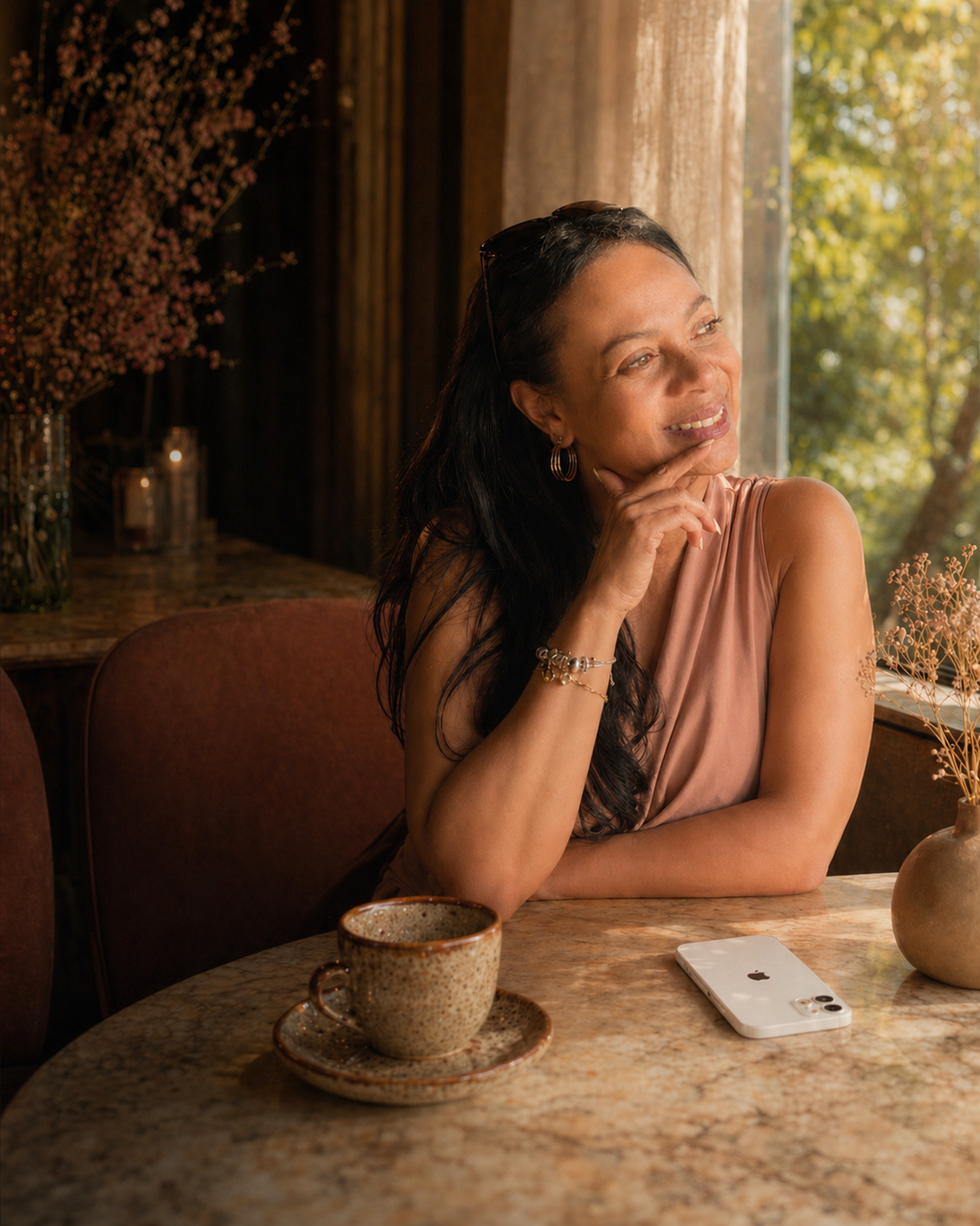 Elegante vrouw in warm natuurlijk licht aan tafel bij een raam, met koffie en telefoon, als beeld bij een blog over aandacht, eigenwaarde en zelfliefde.